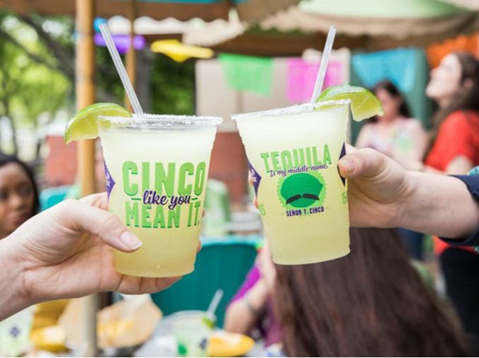 A picture of two hands toasting On The Border margaritas with a bustling street in the background